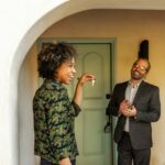 A man and a woman standing outside of a house. The woman excitedly holds up the keys to the front door.