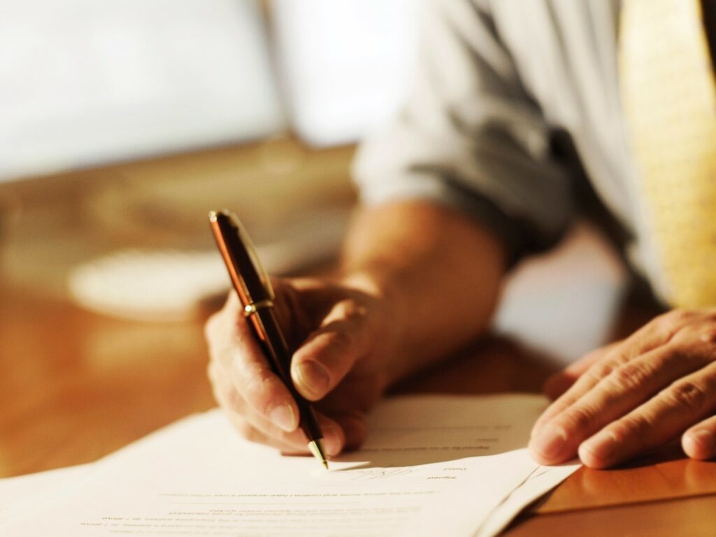 A person in a shirt and tie writes on a document with a pen at a wooden desk.