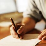 A person in a shirt and tie writes on a document with a pen at a wooden desk.