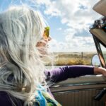 A person with long, gray hair drives a convertible with the top down on a sunny day, wind blowing through their hair.