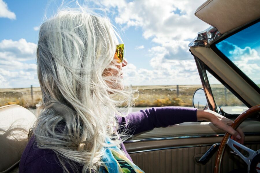 A person with long, gray hair drives a convertible with the top down on a sunny day, wind blowing through their hair.