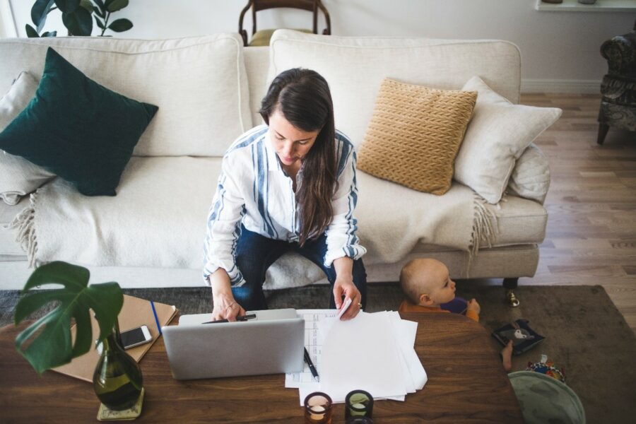 A woman works on a laptop at a coffee table with papers spread out, while a baby sits on the floor beside her in a living room.