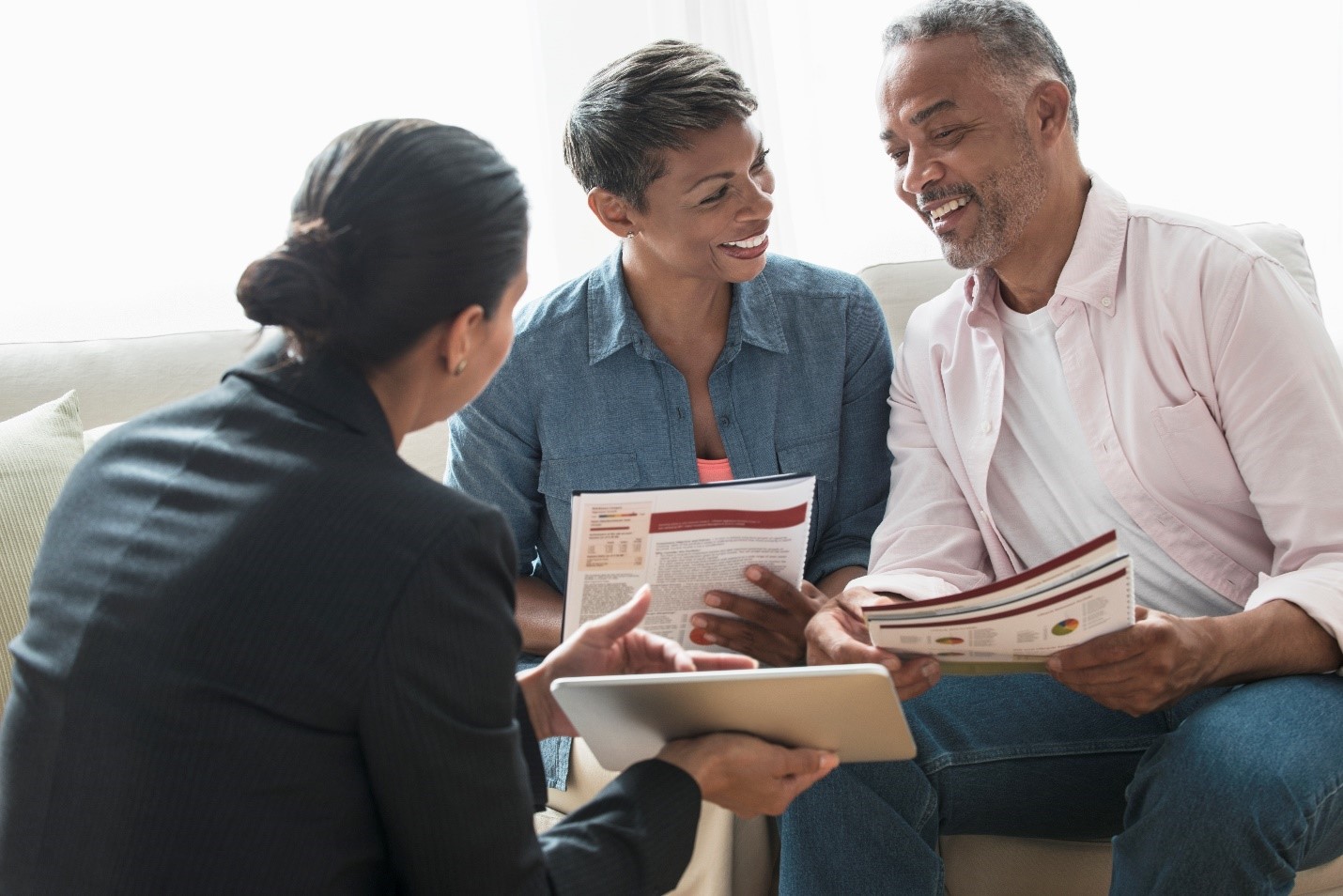  A couple sits on a sofa, smiling and holding documents, as they talk to a person in a suit holding a tablet.