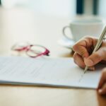 A person is writing on a piece of paper. The scene includes various office supplies such as a pen and scissors on the table, emphasizing the act of handwriting and documentation.