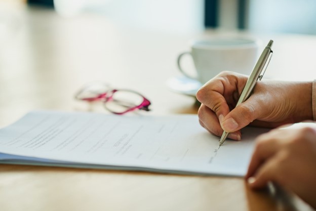 A person is writing on a piece of paper. The scene includes various office supplies such as a pen and scissors on the table, emphasizing the act of handwriting and documentation.