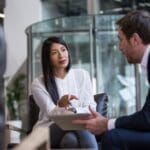 A woman in a white blouse and a man in a suit engage in a serious conversation in a modern office setting, conveying professionalism.