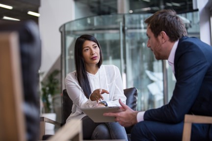 A woman in a white blouse and a man in a suit engage in a serious conversation in a modern office setting, conveying professionalism.